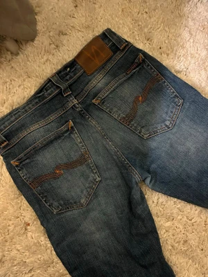 Blå jeans från Nudie Jeans - Säljer ett par klassiska blå jeans från Nudie Jeans med snygg tvätt och kontrastsömmar. Jeansen har fem fickor, normal midja och raka ben. Materialet är slitstark denim i bomull och bakfickorna har Nudies ikoniska broderi.