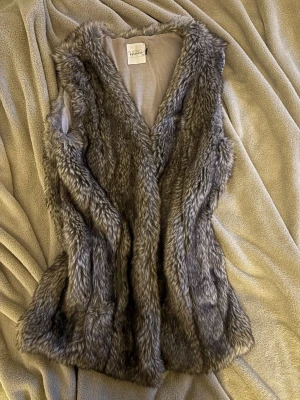 pälsväst  - Snygg grå pälsväst från reserved i fuskpäls. Västen har en mjuk och fluffig känsla, är ärmlös och har en öppen front. Perfekt att slänga över en hoodie eller tröja för en trendig lager-på-lager-look. Storlek S