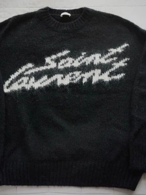  Saint Laurent- mohair tröja - Stickad svart tröja från Saint Laurent med vit grafisk logga över bröstet. Tröjan har rund halsringning, ribbade muddar och är gjord i ett fluffigt material som ger en lyxig känsla. Perfekt för dig som gillar streetstyle och exklusiva märken.