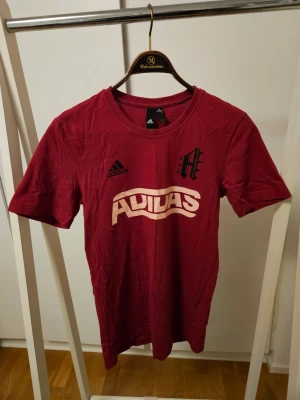 Röd Adidas t-shirt med tryck - Snygg röd t-shirt från Adidas med stort vitt tryck på bröstet och en stor siffra 3 på ryggen. Klassisk rund hals och korta ärmar. Tillverkad i mjuk bomull, perfekt för en sportig och avslappnad stil.