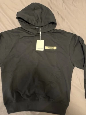 Svart hoodie från Jacquemus - Stilren svart hoodie från Jacquemus med beige tygpatch och svart logotyp på bröstet. Klassisk huva och ribbade muddar vid ärmslut och nederkant. Tillverkad i mjukt bomullsmaterial för en skön och avslappnad look.