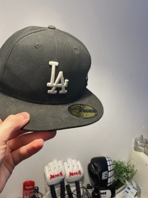 Svart LA New Era 59FIFTY keps - Snygg svart keps från New Era, modell 59FIFTY, med LA-logga broderad i vitt framtill. Klassisk flat brim och strukturerad passform. Tillverkad i bomullsmix och perfekt för dig som gillar streetwear och sportig stil.