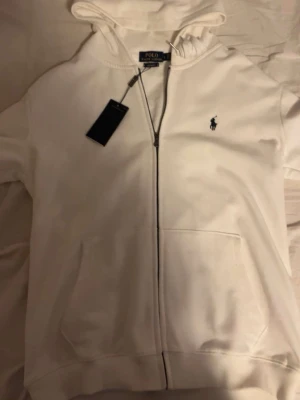 Vit hoodie från Polo Ralph Lauren - Snygg vit hoodie från Polo Ralph Lauren med dragkedja framtill och klassisk huva. Custom fit-modell med två fickor och den ikoniska lilla loggan broderad på bröstet. Perfekt för en clean och stilren look.