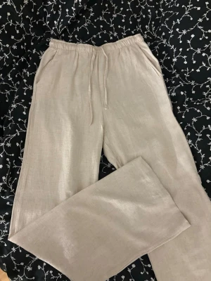 Beiga linnebyxor med dragsko - Jag säljer ett par snygga beiga linnebyxor med avslappnad passform och elastisk midja med dragsko. Byxorna har raka ben och sidofickor. Perfekta för en chill och stilren look till sommaren. Materialet är luftigt linne som känns svalt och skönt.