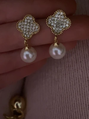 Guldörhängen med pärlor och kristaller - Snygga örhängen i guld med fyrklöverformad topp täckt av små gnistrande kristaller och en hängande vit pärla. Perfekt accessoar för dig som gillar en elegant och trendig look. .