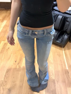 Ljusblå low waist bootcut jeans - Säljer ett par ljusblå low waist jeans med bootcut-modell och dubbla linningar. Jeansen har stora bakfickor med lock och knappar samt klassiska detaljer framtill. Perfekta för en trendig och avslappnad look.