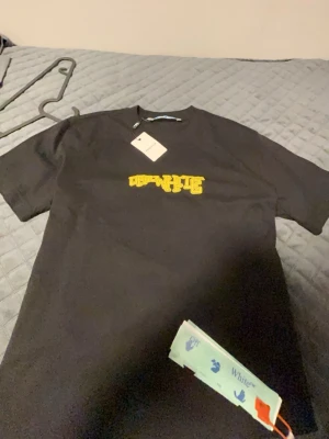 Svart Off-White t-shirt med gul logga - Snygg svart t-shirt från Off-White med gul tryckt logga på bröstet. Klassisk rund hals och korta ärmar. Tillverkad i mjuk bomull för en skön känsla. Perfekt för dig som gillar streetwear och vill sticka ut med en ikonisk design.