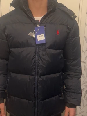 Mörkblå dunjacka från Polo Ralph Lauren - Snygg mörkblå dunjacka från Polo Ralph Lauren med huva och dragkedja framtill. Jackan har det klassiska röda Polo-logotypen broderad på bröstet och är riktigt puffig för maximal värme. Perfekt för kalla vinterdagar och har en modern, sportig look. Priset kan diskuteras 