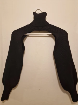 Svart stickad bolero med polokrage - Svart stickad bolero med hög polokrage. Modellen är ribbstickad och har en trendig, minimalistisk look som passar perfekt över en topp eller klänning. Passar även S då det är endast över armarna.