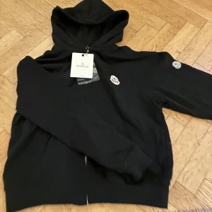 Svart ”Moncler”  hoodie med dragkedja - Snygg svart hoodie från Moncler med dragkedja framtill och klassiska Moncler-loggor på bröstet och ärmen. Stl S - passar dig som är runt 160-170cm