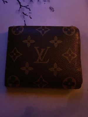 Louis Vuitton plånbok - Säljer en klassisk brun plånbok från Louis Vuitton med det ikoniska monogrammönstret i beige. Plånboken är fyrkantig och tillverkad i tåligt läder med LV-loggan och blommotiv över hela ytan. Perfekt för dig som gillar lyxiga accessoarer.