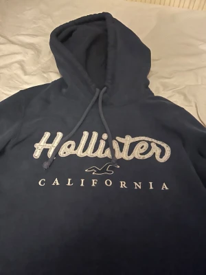 Mörk blå Hollister hoodie - Mörkblå hoodie från Holl CS med stor logga och texten 'California' på bröstet. Klassisk känguruficka framtill och justerbar sopig huva. Perfekt för chill dagar och streetwear-vibe. Mjuk bomullsblandning för extra komfort. Den är inte smutsig på texten det är bara ljuset som gör så den ser ut som det