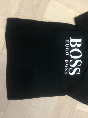Svart BOSS t-shirt med logga - Snygg svart t-shirt från Hugo Boss med stor vit BOSS-logga på bröstet. Klassisk passform och korta ärmar. Tillverkad i mjuk bomull som känns skön mot huden. Perfekt för dig som gillar stilrena och tidlösa plagg.