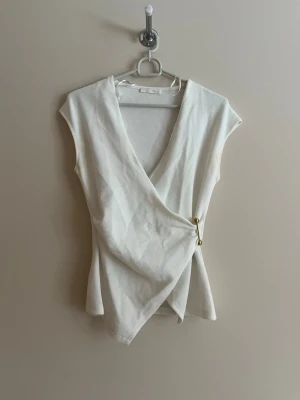 Vit omlottopp med gulddetaljer - Stilren vit omlottopp med v-ringning och två små guldfärgade knappar i sidan. Toppen är ärmlös och har en figurnära passform som framhäver siluetten. Perfekt att matcha med jeans eller kjol för en trendig look.