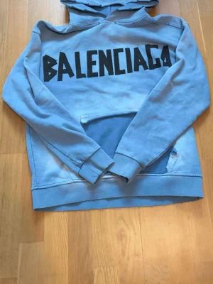Blå Balenciaga hoodie med tryck - Snygg ljusblå hoodie från Balenciaga med stor svart logga på bröstet och ryggen. Klassisk huva och magficka framtill. Oversized passform och mjukt material som känns skönt mot huden. Perfekt för dig som vill sticka ut med streetwear-vibe.
