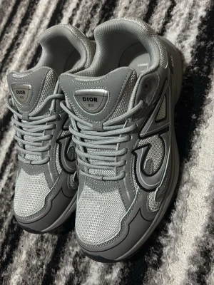 Grå Dior B30 sneakers - Snygga grå Dior B30 sneakers med mesh och läderdetaljer. Skorna har en chunky sula, reflekterande detaljer och tydlig Dior-logga på sidan. Perfekta för dig som gillar exklusiva och trendiga sneakers med sportig vibe.