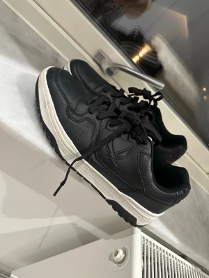 Svarta sneakers med vit sula - Snygga svarta sneakers med vit sula och klassisk design. Skorna har svarta skosnören, diskreta sömmar och en låg profil. Tillverkade i slätt och perforerat konstläder för en clean look. Perfekta för dig som gillar stilrena och tidlösa sneakers.