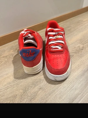 Nike Air Force 1 röda sneakers - Snygga Nike Air Force 1 sneakers i rött läder och mocka med vita detaljer och vita skosnören. Blå detalj med röd 'nike'-brodyr på hälen och klassisk vit sula. Perfekta för dig som vill sticka ut med färg och stil.