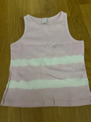 Rosa och vit ribbad linnetopp - Söt ribbad linnetopp i ljusrosa med vita vågiga ränder. Toppen är ärmlös och har en klassisk rund halsringning. Perfekt för varma dagar och enkel att matcha med jeans eller shorts.