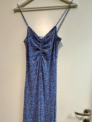 Blommig blå långklänning med smala band - Snygg blå långklänning med smala axelband och blommigt mönster i vitt och rött. Klänningen har rynkade detaljer framtill och en mjuk, följsam passform. Perfekt för sommarens alla tillfällen.