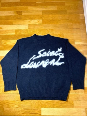 Svart Saint Laurent stickad tröja - Snygg svart stickad tröja från Saint Laurent med vit, fluffig logotyp framtill. Tröjan har rund hals, ribbade muddar och avslappnad passform. Perfekt för dig som gillar streetstyle och exklusiva märken.