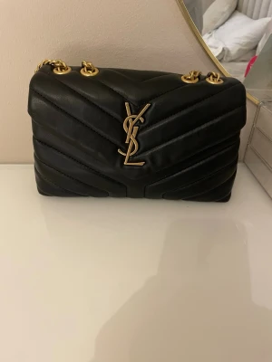 Svart YSL axelväska i läder - Säljer en svart axelväska från YSL i mjukt quiltat läder med guldfärgade kedjor och ikonisk YSL-logga framtill. Väskan har en stilren och lyxig design med snygga detaljer och passar perfekt till dig som vill ha något extra.