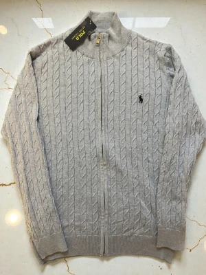 Ljusblå kabelstickad ziptröja Polo Ralph Lauren - Ljusblå kabelstickad tröja med dragkedja från Polo Ralph Lauren. Tröjan har ståkrage, ribbade muddar och den klassiska broderade loggan på bröstet. Perfekt för lager på lager och riktigt snygg till jeans eller chinos.