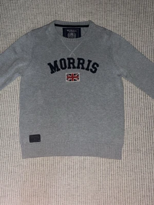 Grå stickad tröja från Morris - Snygg grå stickad tröja från Morris med svart broderad logga och en liten brittisk flagga på bröstet. Tröjan har rund halsringning, långa ärmar och ribbade muddar vid ärmslut och nederkant. Perfekt för en stilren och avslappnad look.