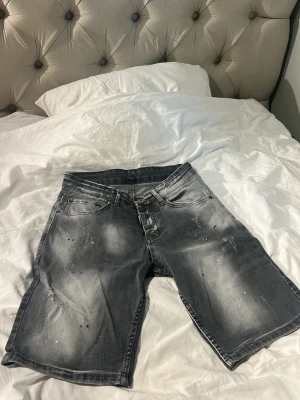 Grå jeansshorts från DSQUARED2 - Säljer ett par grå jeansshorts från DSQUARED2 med slitningar och stänkdetaljer framtill. Shortsen har fem fickor, knappgylf och en patch med DSQ2 och RANTWER på baksidan. Perfekta för en avslappnad streetwear-look.