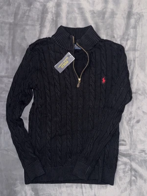 Svart kabelstickad tröja Polo Ralph Lauren - Kabelstickad svart tröja från Polo Ralph Lauren med halv dragkedja och hög krage. Tröjan har det klassiska röda Polo-logot på bröstet och ribbade muddar vid ärmslut och nederkant. Perfekt för lager på lager och snygg till jeans.