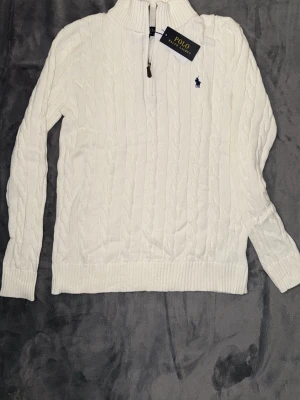 Vit kabelstickad tröja Polo Ralph Lauren - Kabelstickad vit tröja från Polo Ralph Lauren med halv dragkedja och hög krage. Tröjan har ribbade muddar och nederkant samt den klassiska blå logotypen broderad på bröstet. Perfekt för dig som gillar stilrena och tidlösa plagg.