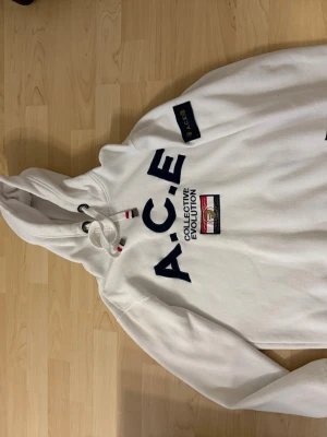 Vit hoodie från WHO's A.C.E - Snygg vit hoodie från WHO's A.C.E med stor logga och texten 'COLLECTIVE EVOLUTION' på bröstet. Hoodien har huva med dragsko och detaljer i rött och vitt. Mjuk insida och patch på ärmen. Perfekt för en chill och trendig look.
