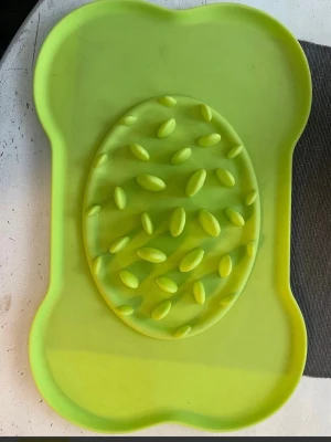 Grön slickmatta för hund - Säljer en grön slickmatta i silikon, formad som ett ben med ett ovalt mönstrat område i mitten. Perfekt för att aktivera hunden vid mat eller godis, el att få den äta långsammare.  Mattan har upphöjda detaljer som gör det svårare för hunden att slicka upp allt direkt.Lägg mjukmat i mitten och smeta ut, in i frysen och sen låt hunden slicka i sig maten. 