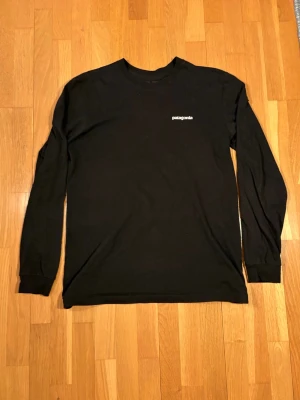 Svart långärmad Patagonia t-shirt - Säljer en svart långärmad t-shirt från Patagonia med liten logga fram och stor färgglad logga med bergmotiv på ryggen. Klassisk rund hals och mjukt material som känns skönt mot huden. Perfekt för dig som gillar stilrena och ikoniska plagg.