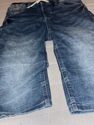 Blå jeansshorts med snörning - Säljer ett par klassiska blå jeansshorts från L.O.G.G. med vit snörning i midjan. Shortsen har normal passform och är tillverkade i slitstarkt denimtyg. Perfekta för en avslappnad stil under varmare dagar.
