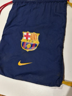 Blå  Barcelona gympåse Nike - Mörkblå gympåse med FC Barcelona-logga och gul Nike-swoosh på framsidan. Påsen har röda och gula snören som axelband och är tillverkad i ett lätt syntetmaterial. Perfekt för träning eller skola för dig som gillar fotboll och Barca.