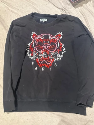 Svart sweatshirt med tiger från Kenzo - Cool svart sweatshirt från Kenzo med ikonisk broderad tiger i rött, vitt och rosa på bröstet samt texten 'KENZO PARIS'. Tröjan har rund halsringning och långa ärmar. Perfekt för dig som gillar streetwear och vill sticka ut. 250kr tills helgen