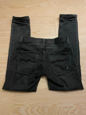 Svarta slim fit jeans från Nudie Jeans - Säljer ett par svarta slim fit jeans från Nudie Jeans med klassiska bakfickor och diskret logga på ena fickan. Jeansen har fem fickor, normal midja och är tillverkade i denimtyg för skön passform. Passformen heter grim Tim