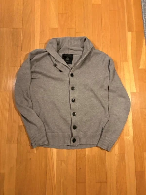 Grå cardigan från Jack & Jones - Snygg grå kofta från Jack & Jones Premium med knäppning framtill. Tillverkad i mjukt material, har ribbade muddar vid ärmslut och nederkant samt stora svarta knappar. Perfekt för en avslappnad och stilren look.