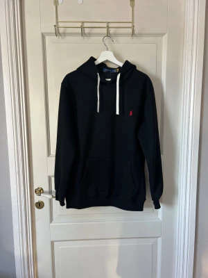 Svart hoodie från Polo Ralph Lauren - Svart hoodie från Polo Ralph Lauren med vit dragsko i huvan och röd broderad logga på bröstet. Klassisk känguruficka framtill och ribbade muddar vid ärmslut och nederkant. Perfekt för en avslappnad och stilren look. Strl XL men snarare en M.