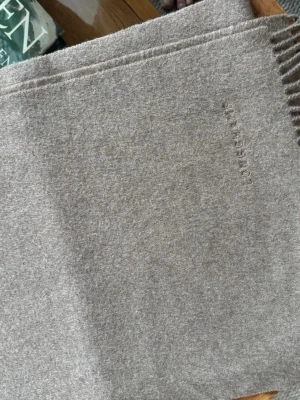 Halsduk i 100% kashmir från Longchamp - Stilren grå halsduk från Longchamp i mjuk kashmir. Halsduken har en enkel design med diskret broderad logga och fransar i  änden. Perfekt för att hålla värmen och addera en klassisk touch till din outfit.