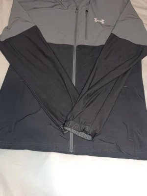 Grå och svart hoodie från Under Armour - Sportig hoodie från Under Armour i grått och svart med dragkedja framtill och två sidofickor. Långärmad modell med logga på bröstet och tryck på ärmsluten. Perfekt för träning eller chill dagar.