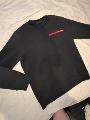 Svart prada tröja  - Snygg svart sweatshirt från Prada med rund halsringning och en röd logodetalj på bröstet. Tröjan har en clean och stilren look utan huva och är tillverkad i ett mjukt material som känns skönt mot huden. Perfekt för dig som gillar exklusiva streetwear-plagg.