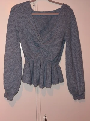 Blå  stickad topp med twistad front - Säljer en blå stickad topp med twistad detalj framtill och peplum nedtill. Tröjan har långa puffärmar och djup v-ringning. Perfekt för dig som gillar trendiga och bekväma plagg i mjukt material. Storleken känns som M 