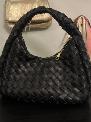 Svart flätad handväska i skinnimitation - Trendig svart handväska med flätad design och rundad form. Väskan har ett rejält handtag och stängs med guldfärgad dragkedja. Tillverkad i skinnimitation som ger en lyxig känsla. Perfekt accessoar för att lyfta din outfit.