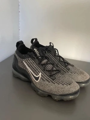 Nike Vapormax Flyknit svarta sneakers - Nike Vapormax Flyknit sneakers i svart och grått med vit Swoosh på sidan. Ovandelen är stickad mesh för extra andningsförmåga och sulan har synliga luftbubblor för maxad komfort. Skorna har snörning och en sportig, modern look.