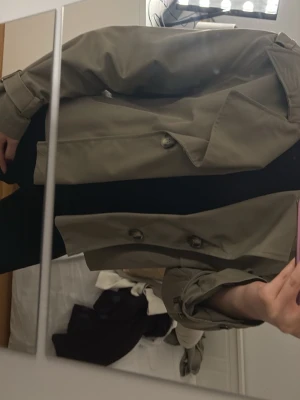 Kort beige trenchcoat med stora slag - Snygg kort trenchcoat i beige med dubbelknäppning och stora slag. Jackan har axelklaffar, bälte vid ärmsluten och en klassisk krage. Tillverkad i ett slätt tyg som ger en stilren look. Perfekt för dig som vill ha en trendig och tidlös jacka.