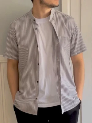Randig kortärmad skjorta från Ovin - Snygg kortärmad skjorta från Ovin med smala vita och grå ränder. Skjortan har ståkrage, bröstficka och knappar framtill. Perfekt för en avslappnad men stilren look. Tillverkad i mjuk bomull som känns skön mot huden.