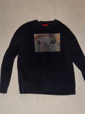 Svart sweatshirt från HUGO - Cool svart sweatshirt från HUGO med stor logga i silver och grått framtill. Tröjan har rund halsringning och ribbade muddar vid ärmslut och nederkant. Perfekt för dig som gillar streetwear och vill ha en stilren look.