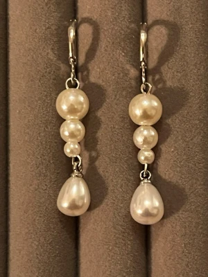 Silverörhängen med vita pärlor - Säljer ett par eleganta örhängen i silverfärg med vita pärlor i olika storlekar. Örhängena har droppformad pärla längst ner och klassisk krokstängning. Perfekta för dig som gillar stilrena accessoarer med en twist.